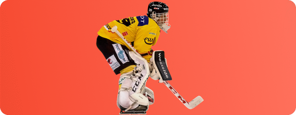Ice hockey goalie in yellow jersey and protective gear holding a stick, isolated on an orange background